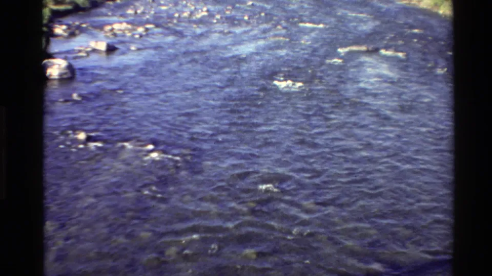 Archival 8mm view of the same rocky river, with the water occupying most of the frame and grassy shoreline only partly visible.