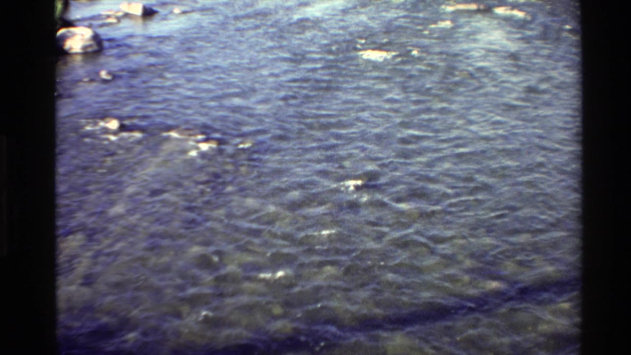 1980 Western USA 8mm frame showing clear shallow water, submerged stones, soft grain, and dark film gate edges.