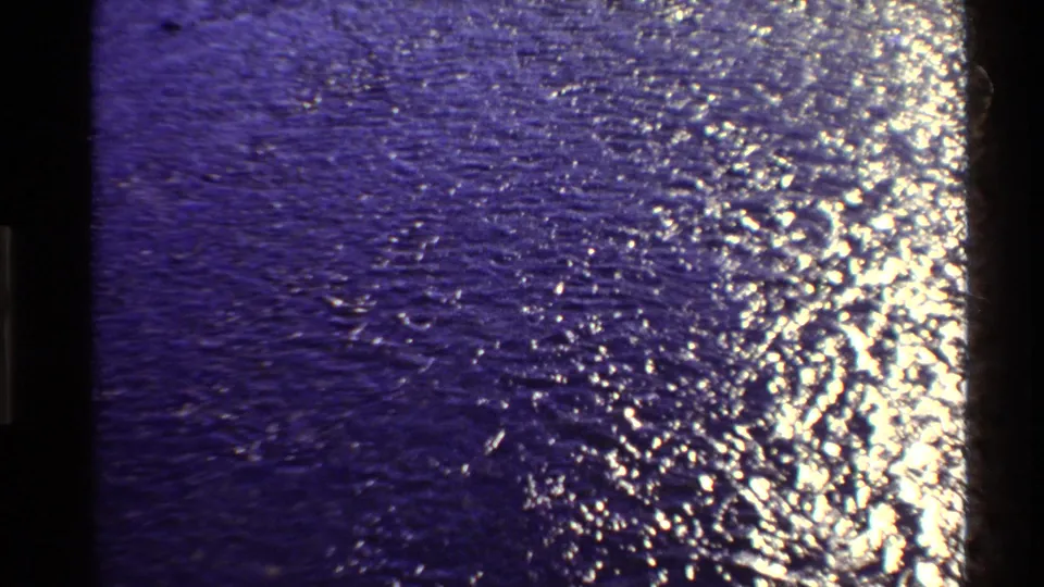 1980 8mm home-movie frame showing blue-purple rippling water, bright reflections, grain, and frame-edge darkness.