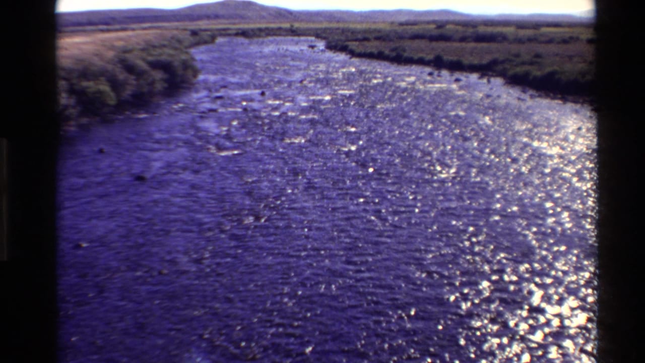 Archival 8mm view of a river running through open Western U.S. terrain in 1980, with wind ripples and bright glare on the water.