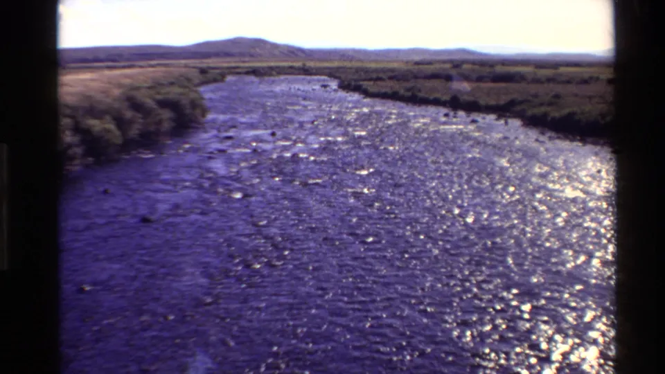 1980 home-movie frame of a reflective river channel bordered by low vegetation and open fields in the Western United States.