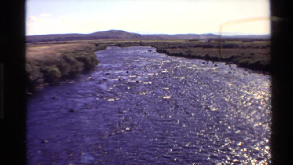 Western U.S. 8mm landscape frame from 1980 showing a sunlit river, open field margins, and a low hill under a pale sky.