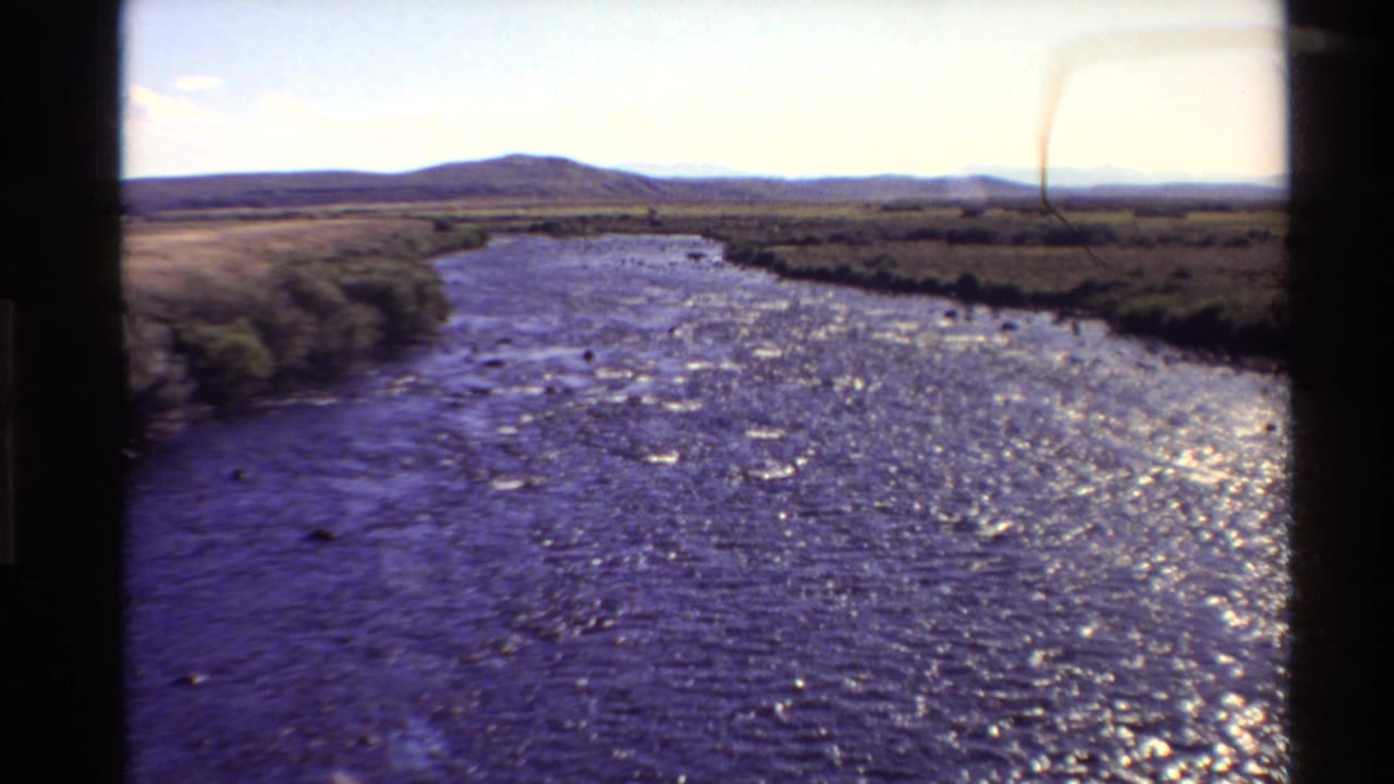 1980 Western U.S. 8mm frame showing a sunlit river, green riverside vegetation, distant hills, and visible film gate edges.