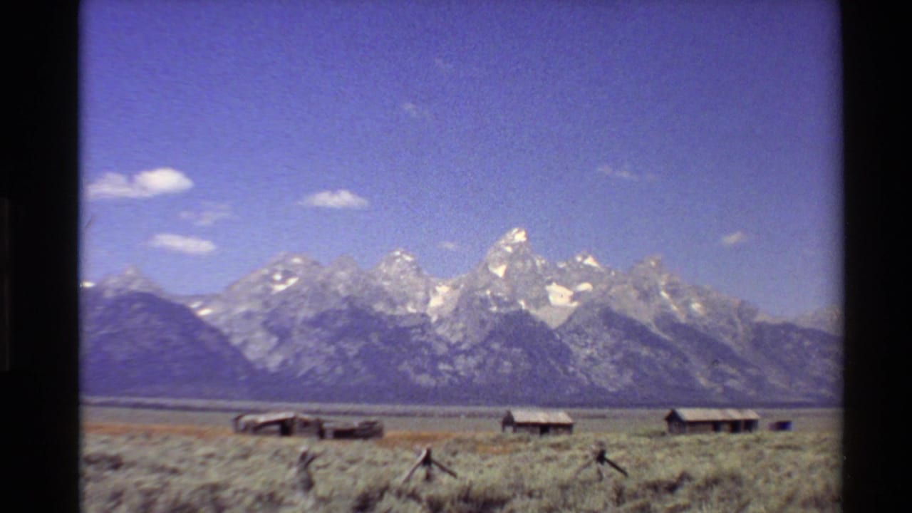 1980 Western U.S. 8mm frame of snow-capped mountains rising beyond an open plain with weathered wooden cabins below.