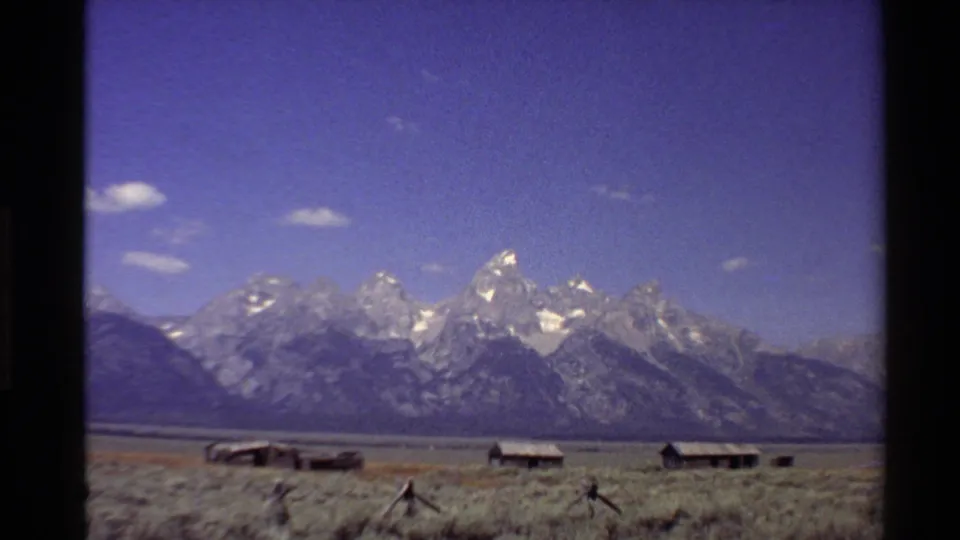 1980 Western U.S. 8mm frame showing a mountain range, open scrubland, low wooden cabins, and small clouds in a clear sky.