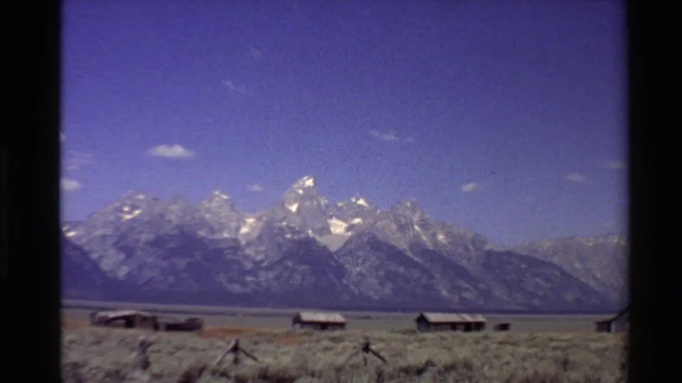 1980 Western U.S. 8mm frame with snowy mountain peaks, a broad ridgeline, open plain, cabins, and visible film softness.
