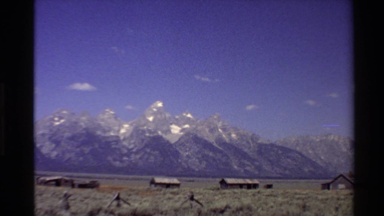 1980 Western U.S. 8mm frame showing snow-capped peaks beyond dry grassland, rustic cabins, and a low wooden fence.