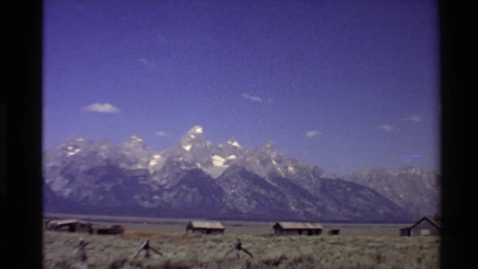 1980 Western U.S. 8mm frame showing snow-capped peaks beyond dry grassland, rustic cabins, and a low wooden fence.