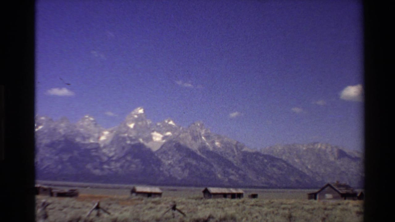 Wide mountain landscape with cabins, dry plain, fence posts, and blue sky.