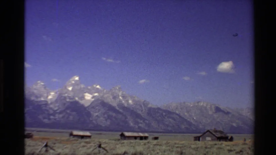 1980 Western U.S. 8mm frame with snowy peaks, a long ridgeline, rustic cabins, and dry foreground grass.