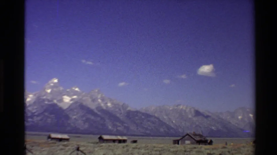 1980 Western U.S. 8mm frame showing cabin exteriors on a dry plain with mountains and scattered clouds beyond.