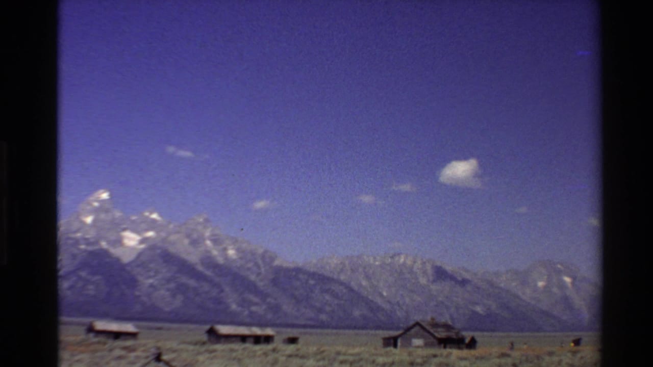 Mountain range behind open fields and small farm buildings under a blue sky.