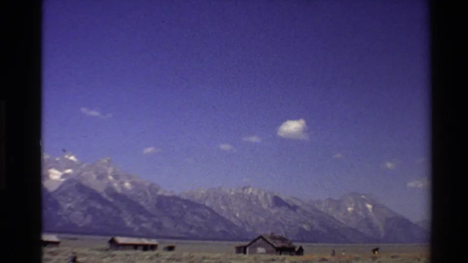 Wide 8mm view of farm buildings and open land set against a Western USA mountain range in bright sun.