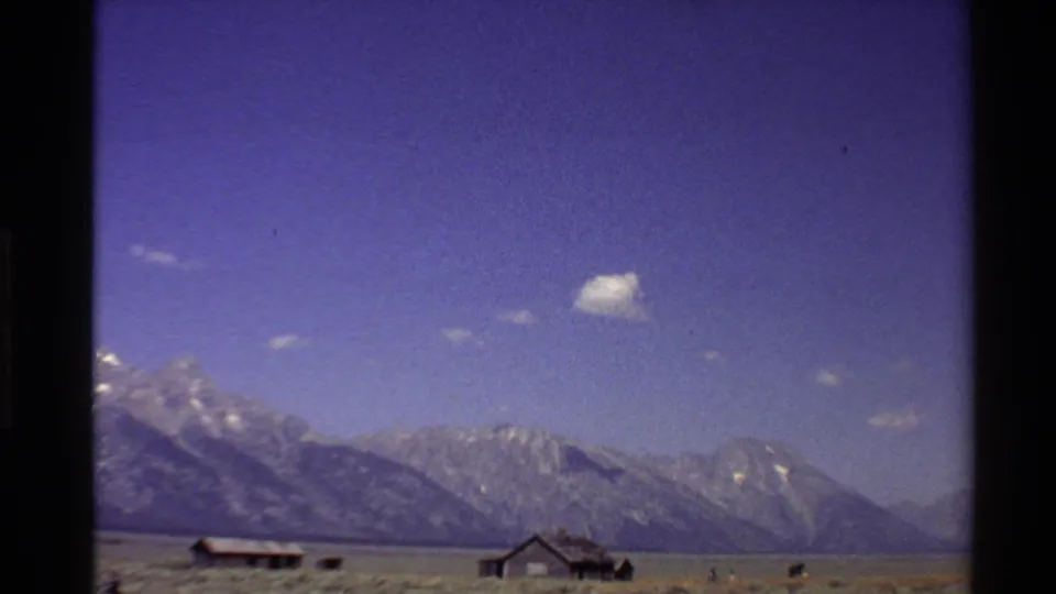 Home-movie frame from the 1980 Western USA clip with low farm structures, open field, and distant mountains.