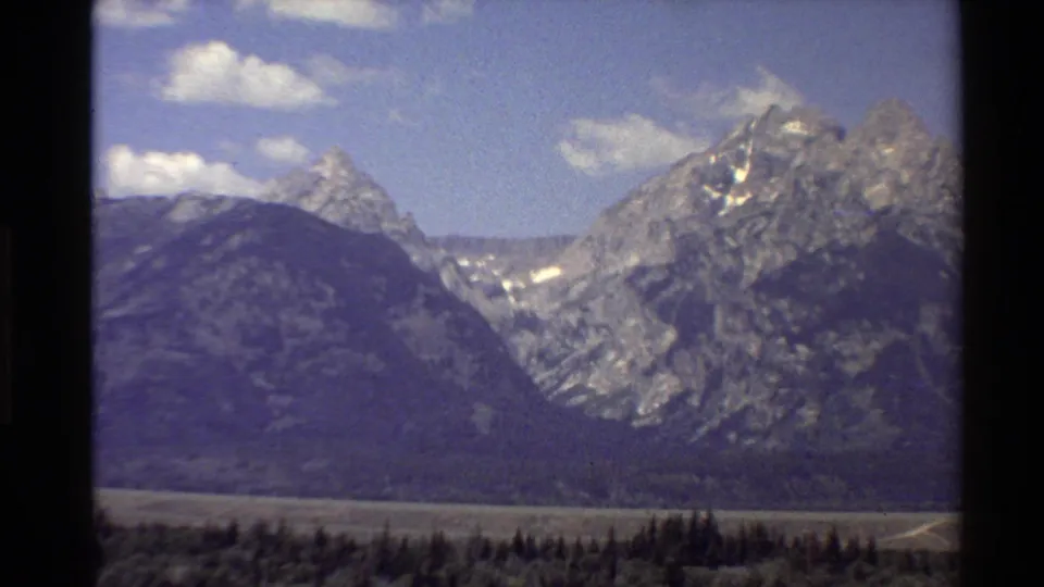 Closer 8mm view of dramatic Western USA mountain peaks with forested foothills and bright clouds overhead.