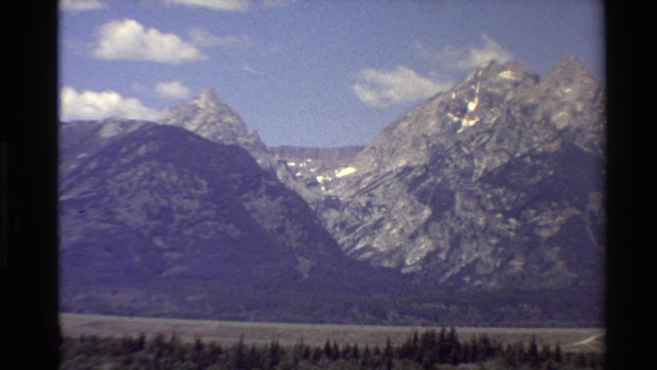 1980 Western USA 8mm frame showing rugged mountain peaks, a shadowed valley, forested foreground, and scattered clouds.