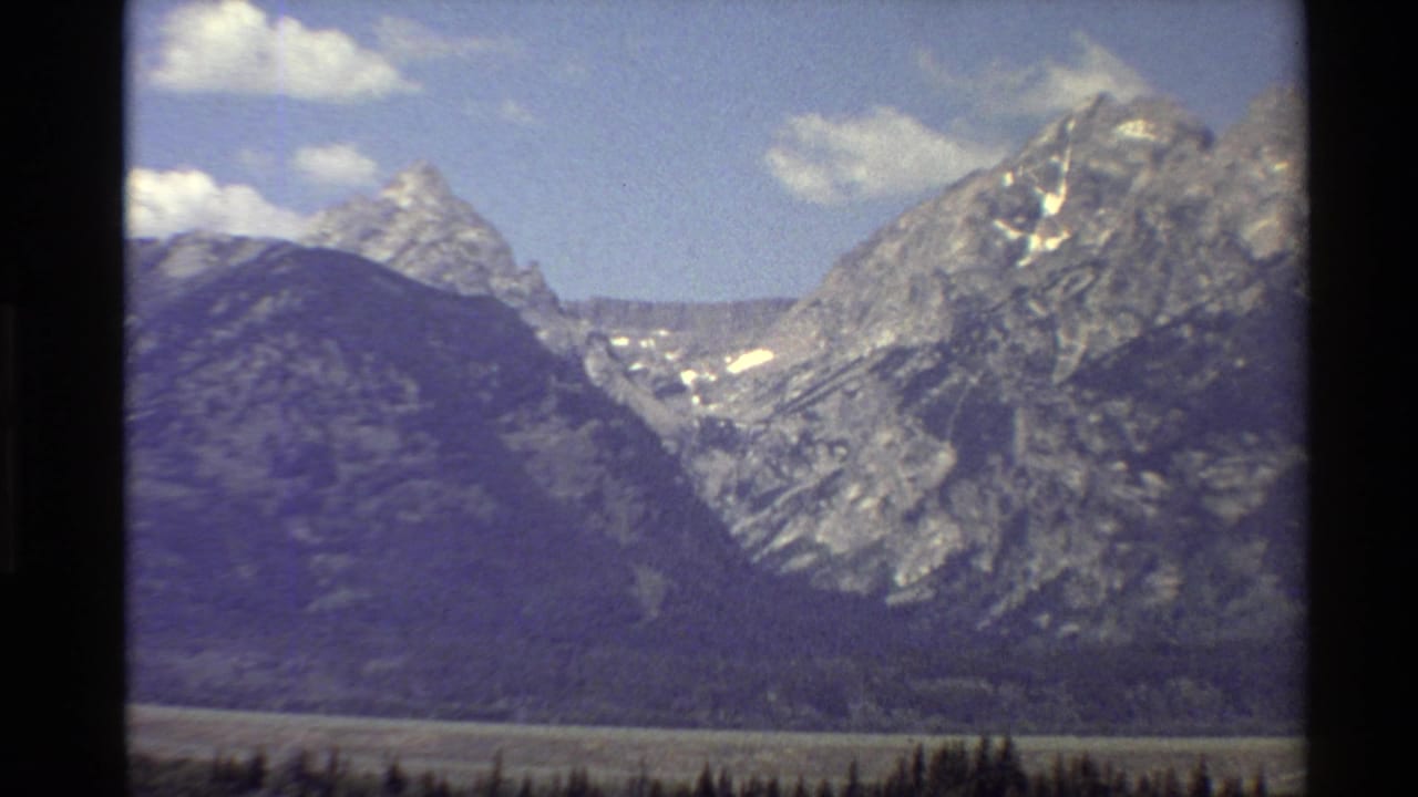 1980 Western USA 8mm frame with a wide mountain panorama, snow patches on rocky slopes, and dark forest at the base.