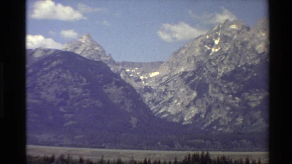 1980 Western USA 8mm frame with a wide mountain panorama, snow patches on rocky slopes, and dark forest at the base.