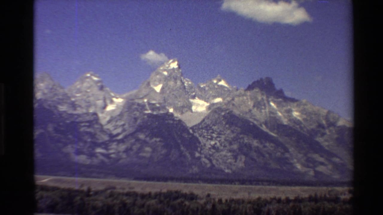 Wide mountain landscape with snow patches, rocky ridges, blue sky, and a small cloud.