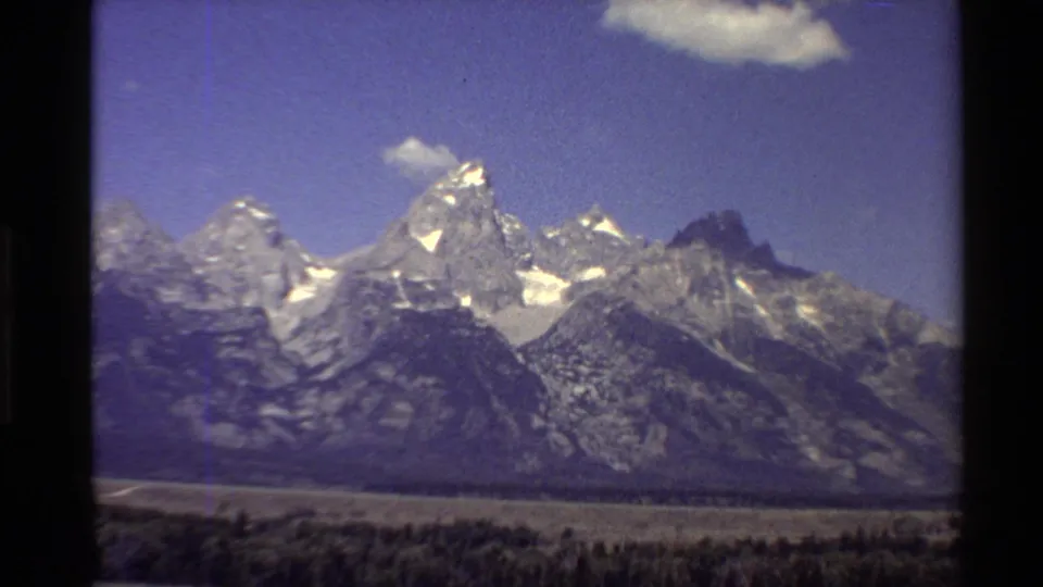 Western U.S. 1980 home-movie frame of a rugged snow-capped mountain range under clear blue sky, with open land below.