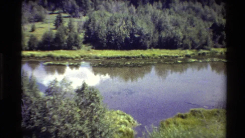 1980 Western USA 8mm frame with the camera closer on a lake, far shore vegetation, shrubs, and forested background.