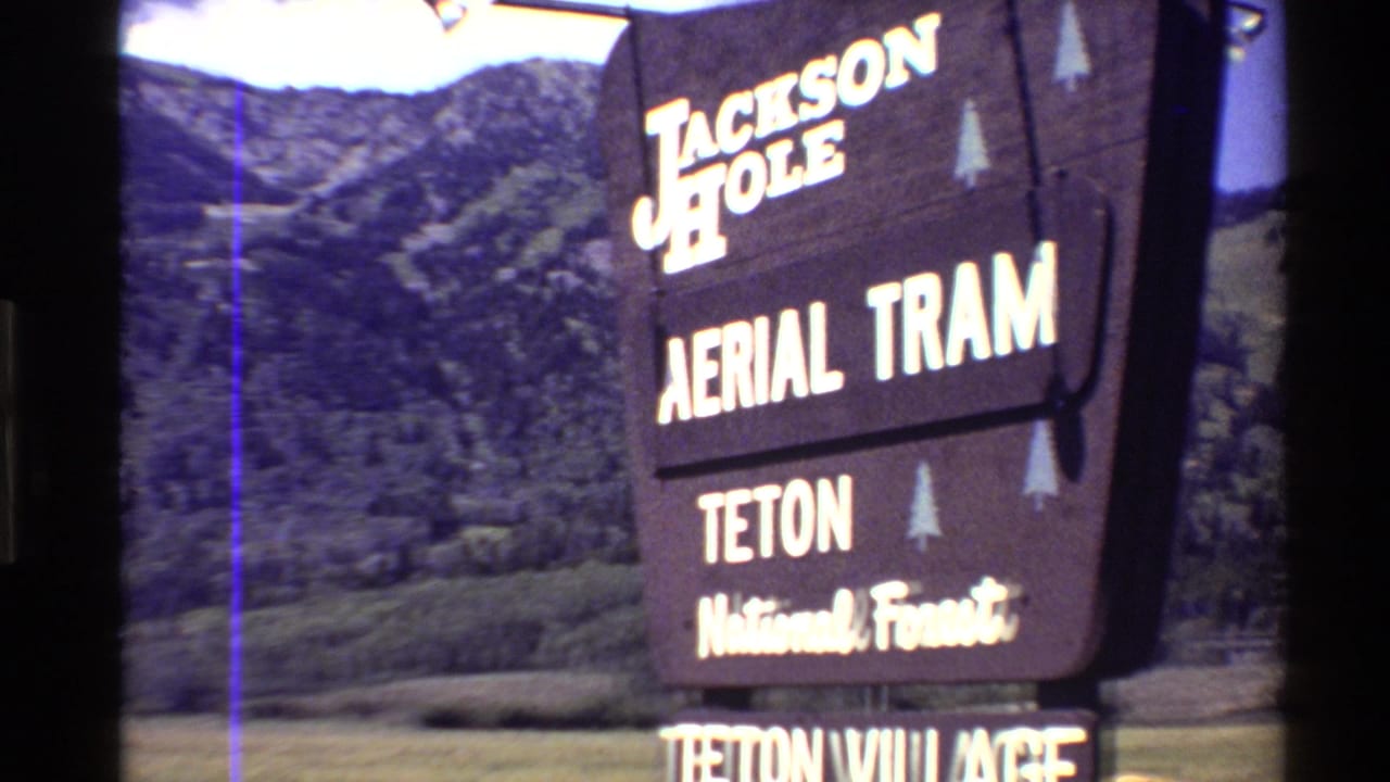 1980s 8mm frame showing a Jackson Hole Aerial Tram and Teton National Forest sign with summer mountain slopes behind it.