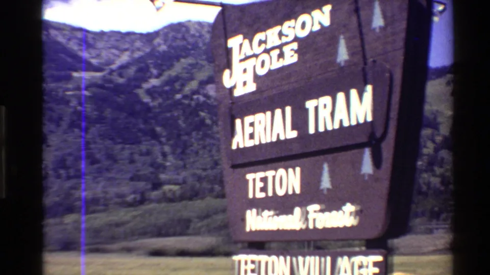 1980s 8mm frame showing a Jackson Hole Aerial Tram and Teton National Forest sign with summer mountain slopes behind it.