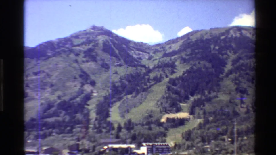 Western U.S. resort mountain in summer, with dry ski runs, trees, and low buildings at the base.