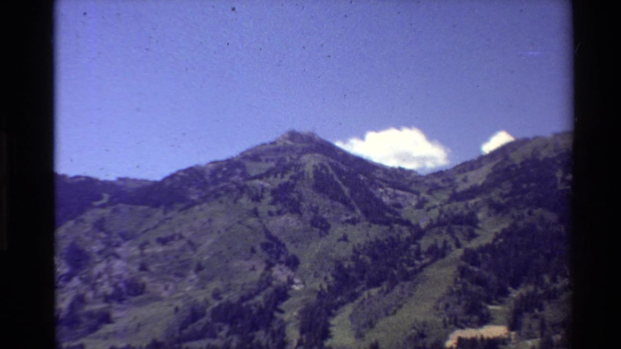 A handheld 8mm view of green mountain slopes and tree-covered ridges in the Western USA, with a bright blue sky and visible film border shadows.