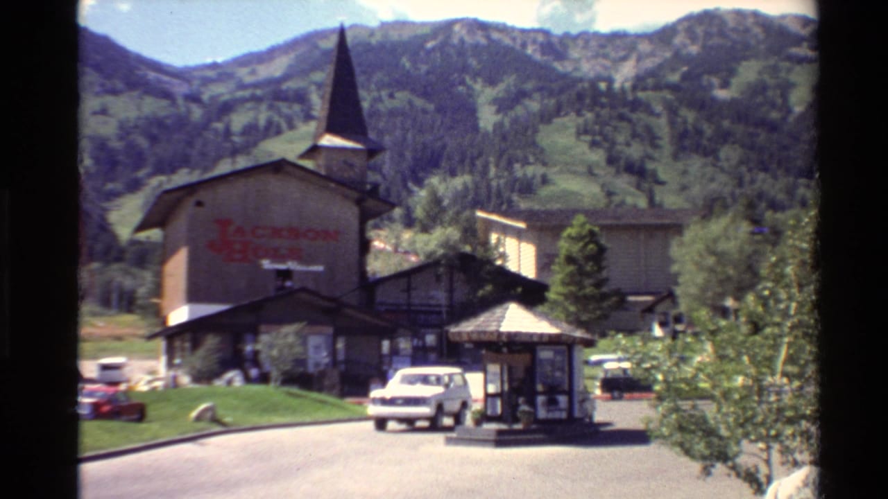 Rustic Jackson Hole building with steeple, road, kiosk, parked cars, and mountains behind.