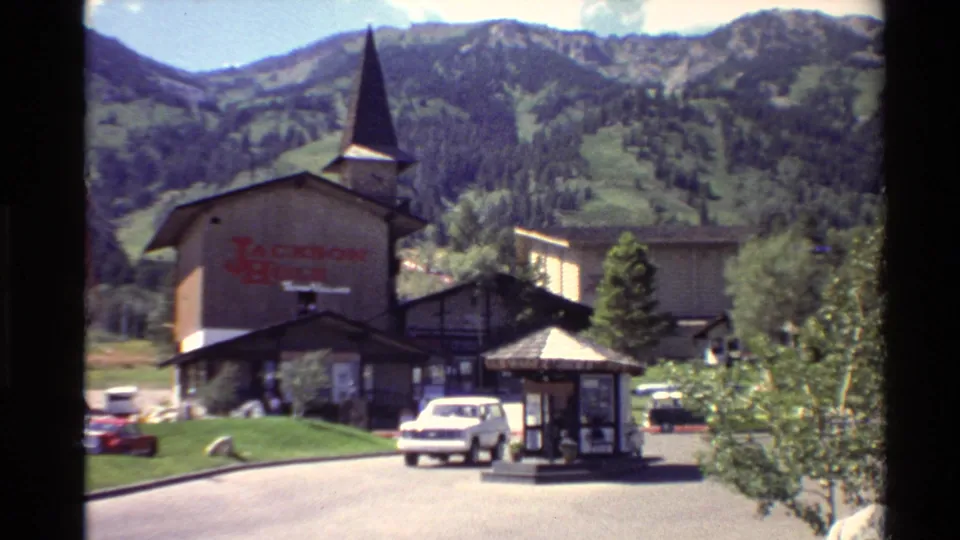 A rustic Jackson Hole Teton Village building with a pointed steeple stands below forested mountains as a white vehicle approaches near a small roadside kiosk.