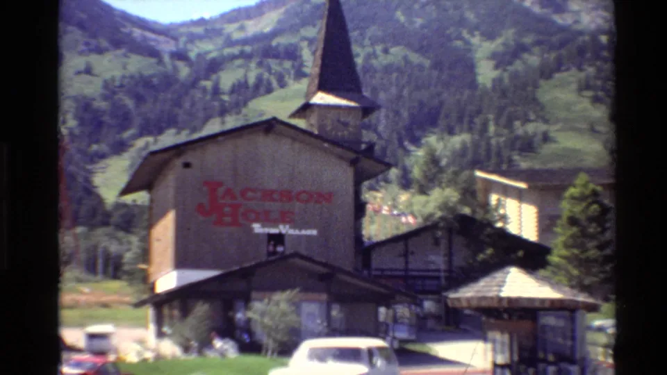 The camera frames the Jackson Hole Teton Village building more closely, showing red signage, a steeple, parked cars, a white vehicle, and alpine slopes behind.