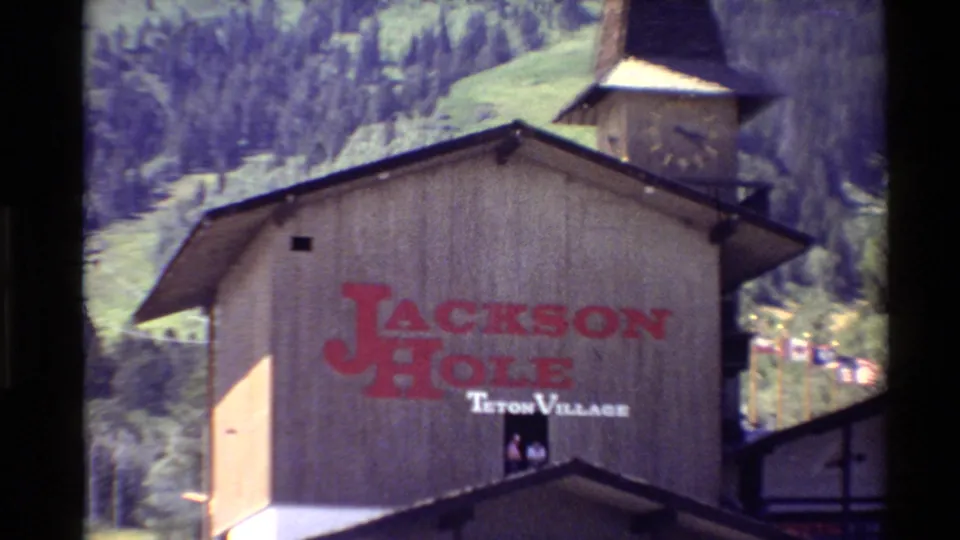 A close 8mm frame shows the red Jackson Hole name and Teton Village text on a rustic building, with its steeple and green mountain slopes partly visible behind.