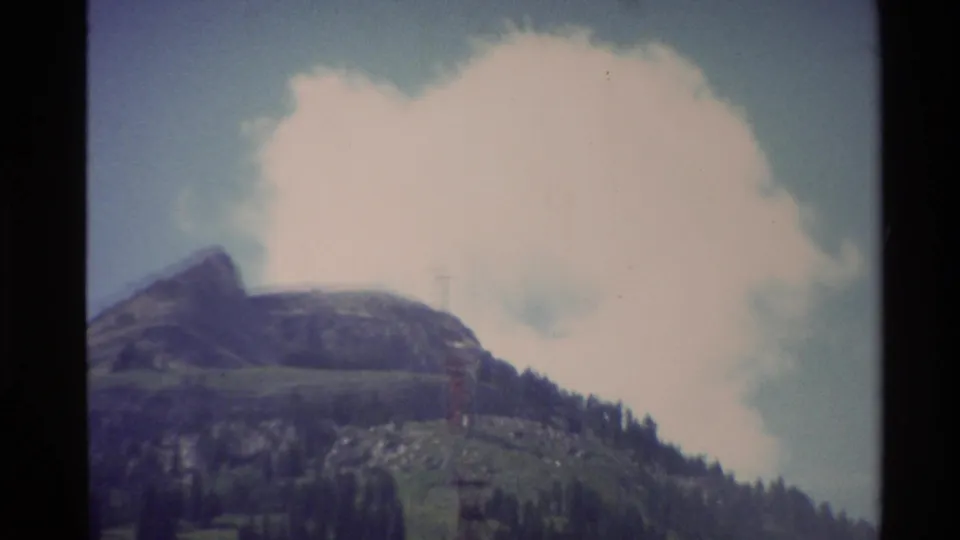 Vintage 8mm frame of a mountain peak, forested hillside, large cloud, and red cable car tower in the Western USA.