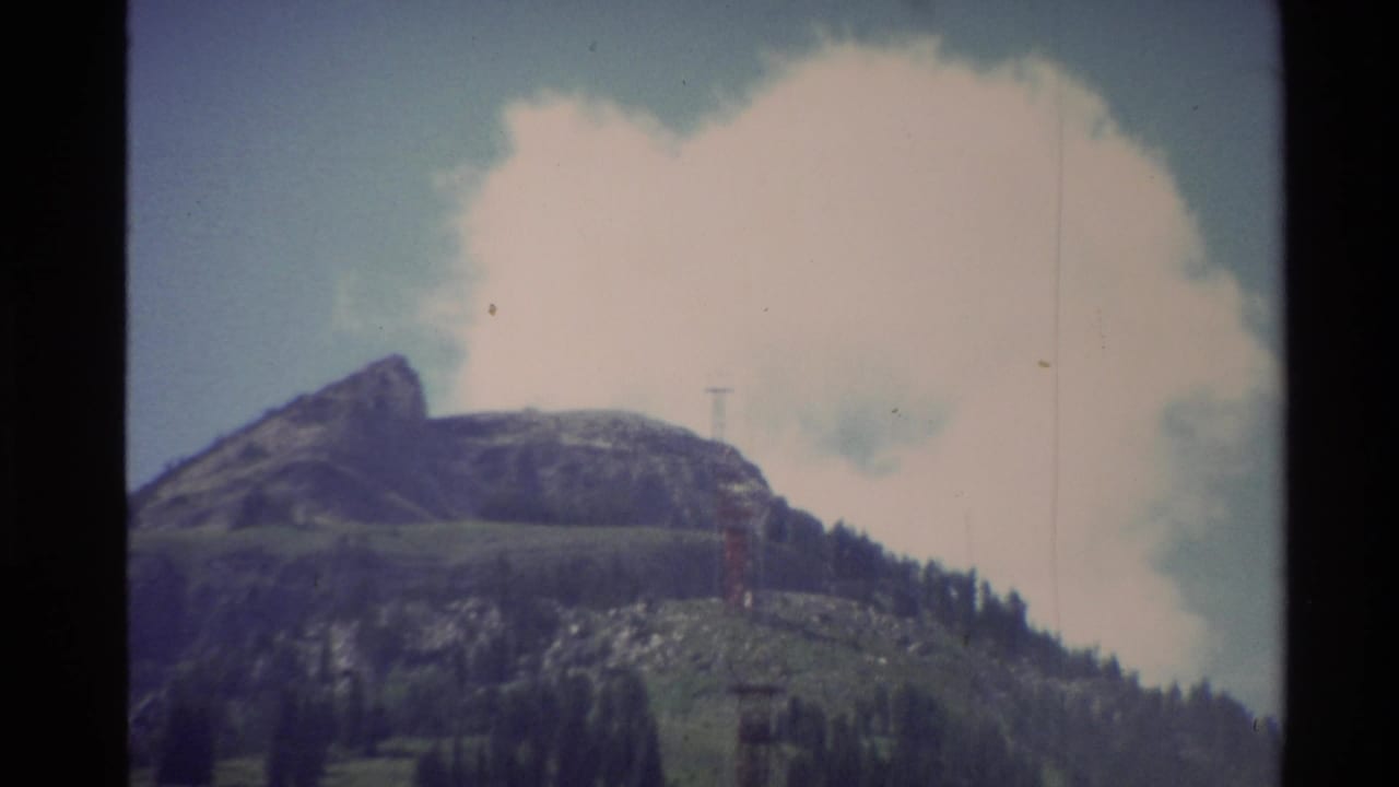 1980s 8mm frame of a Western U.S. mountainside with a rugged peak, forested lower slope, bright cloud, and small vertical tower or lift structure.