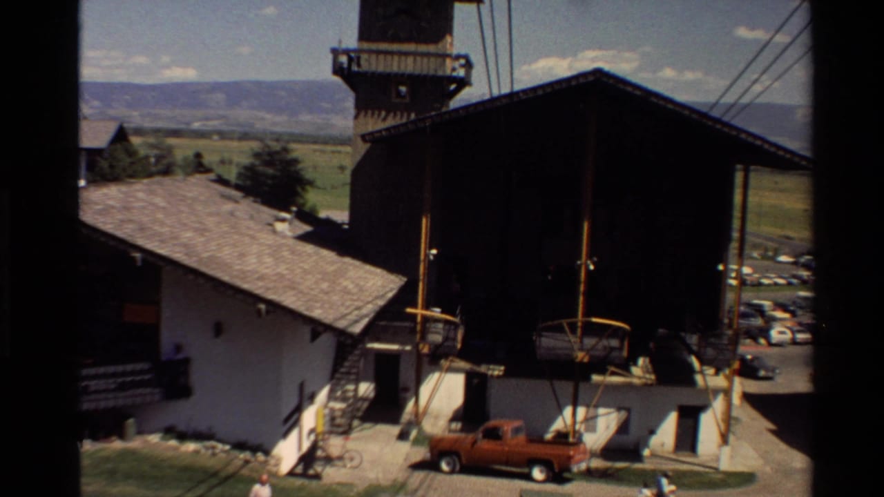 Wooden towered building beside lift equipment, parked cars, valley hills, and an orange pickup truck.