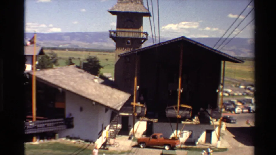1980s 8mm view from a moving gondola looking down at a mountain lift building, cables, parking area, valley floor, and orange pickup truck.