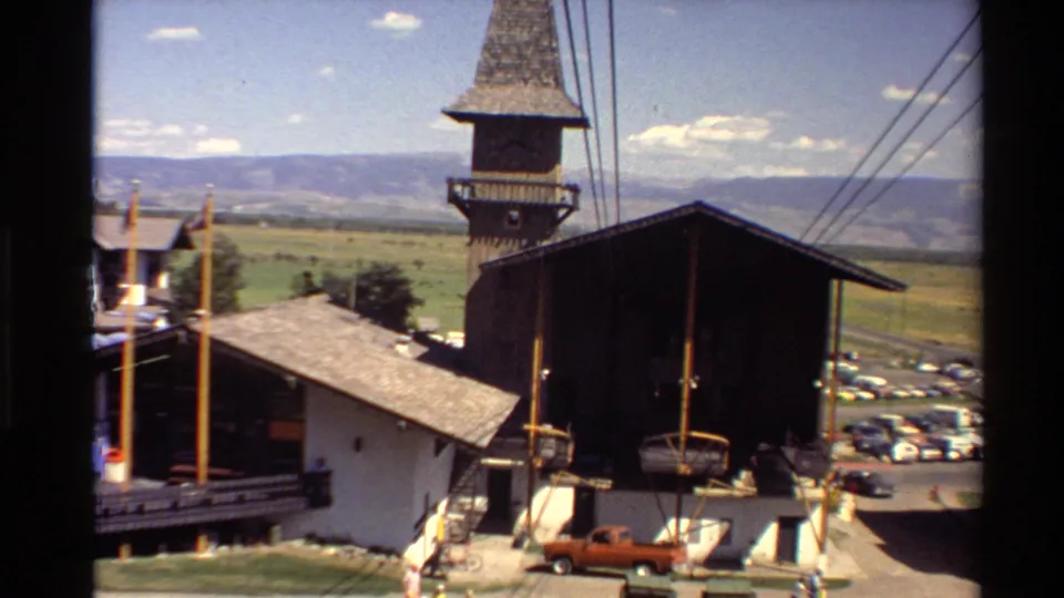 Western U.S. 1980s 8mm frame showing a gondola or lift approach past a towered wooden building, chair equipment, parked cars, and a broad valley.