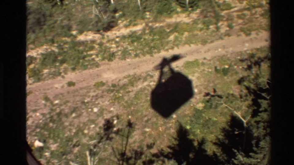The cable car shadow falls across a sunlit slope with scattered trees, brush, and a dirt road, captured in soft 8mm color.