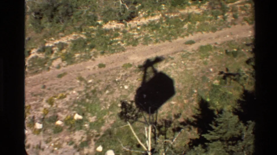 From above, the gondola shadow hangs over trees and scrub beside a dirt track, with home-movie grain and soft exposure visible.