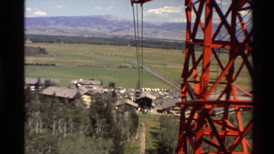 A red cable car tower and hanging cables frame a Western U.S. mountain-town view with buildings, parked cars, fields, and distant mountains.