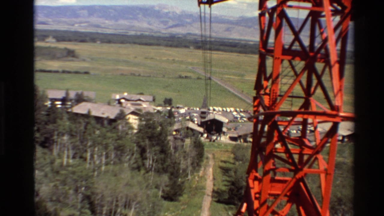 1980 8mm view from a cable car or lift, with a red steel support tower in front of trees, small buildings, open fields, and distant hills.