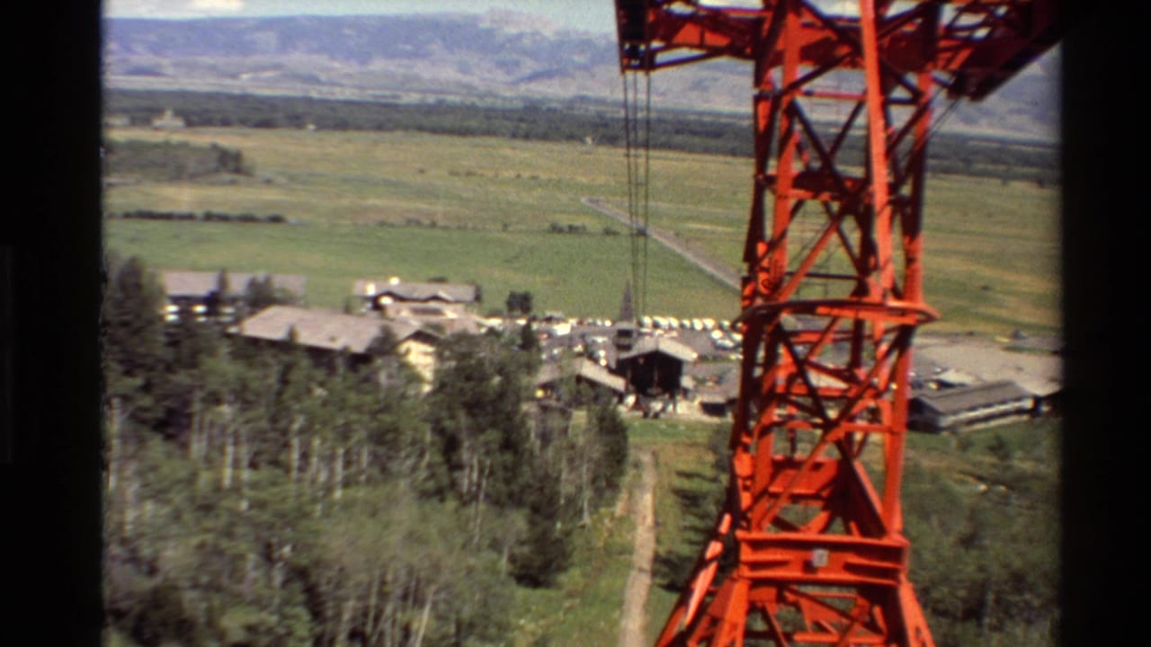 Red lift structure and cables overlook trees, clustered buildings, fields, and distant mountains.