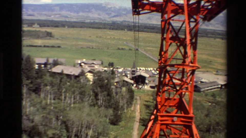Western USA home-movie frame showing a red lift support structure and cables above pine trees, town buildings, parking areas, and distant hills.