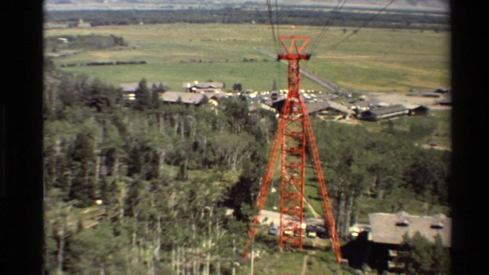 A centered red cable-lift tower rises from wooded ground, with rooftops, parked vehicles or lots, open fields, and hazy hills beyond.