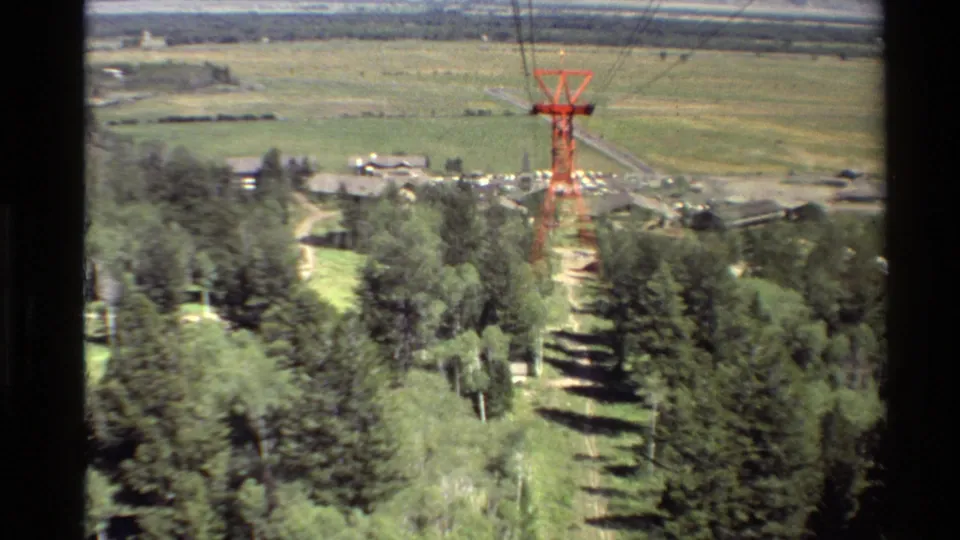 End frame from the moving lift shows the red tower farther away over dense trees, with small buildings, open fields, and distant hills in soft 8mm color.