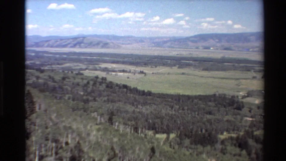 1980 Western United States landscape frame showing a broad valley, forested foreground, low mountains, and blue sky.