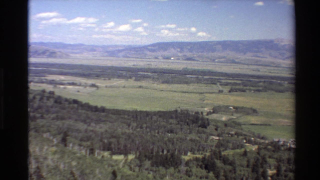 1980s 8mm view from above a Western USA valley, showing green fields, pine forest, distant mountains, and scattered clouds.