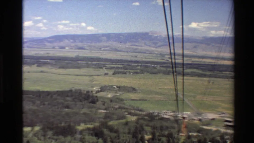 The moving 8mm camera looks across open fields and forest toward a mountain range, with lift cables visible in the foreground.