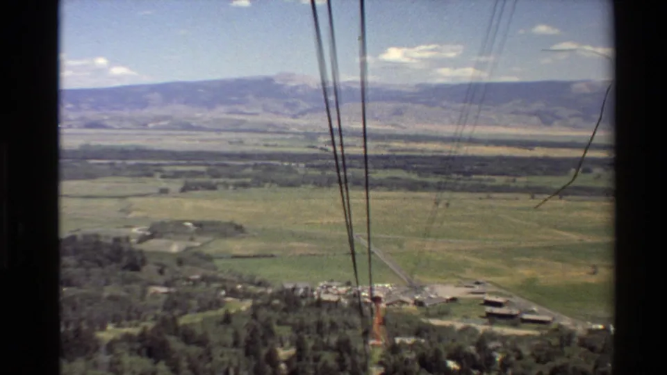 Aerial 8mm view of a Western USA valley with lift cables in front, fields and trees below, and a small building cluster near the lower edge.